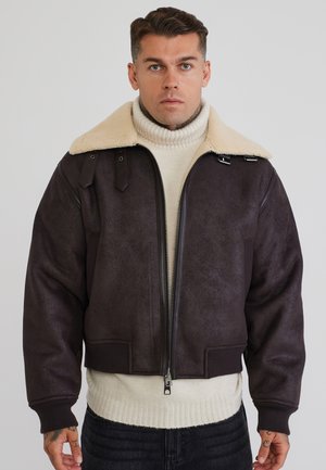 Uomo che indossa una giacca in shearling marrone scuro con ampio colletto sopra un maglione dolcevita bianco, in piedi davanti a uno sfondo chiaro e uniforme.