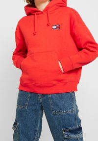 Red hooded sweatshirt with adjustable drawstrings, front pocket, ribbed cuffs, and hem. Features a small black and white logo patch.