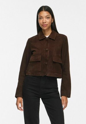 Femme portant une veste marron foncé boutonnée avec de grandes poches à l'avant et un pantalon noir, debout devant un fond blanc uni.