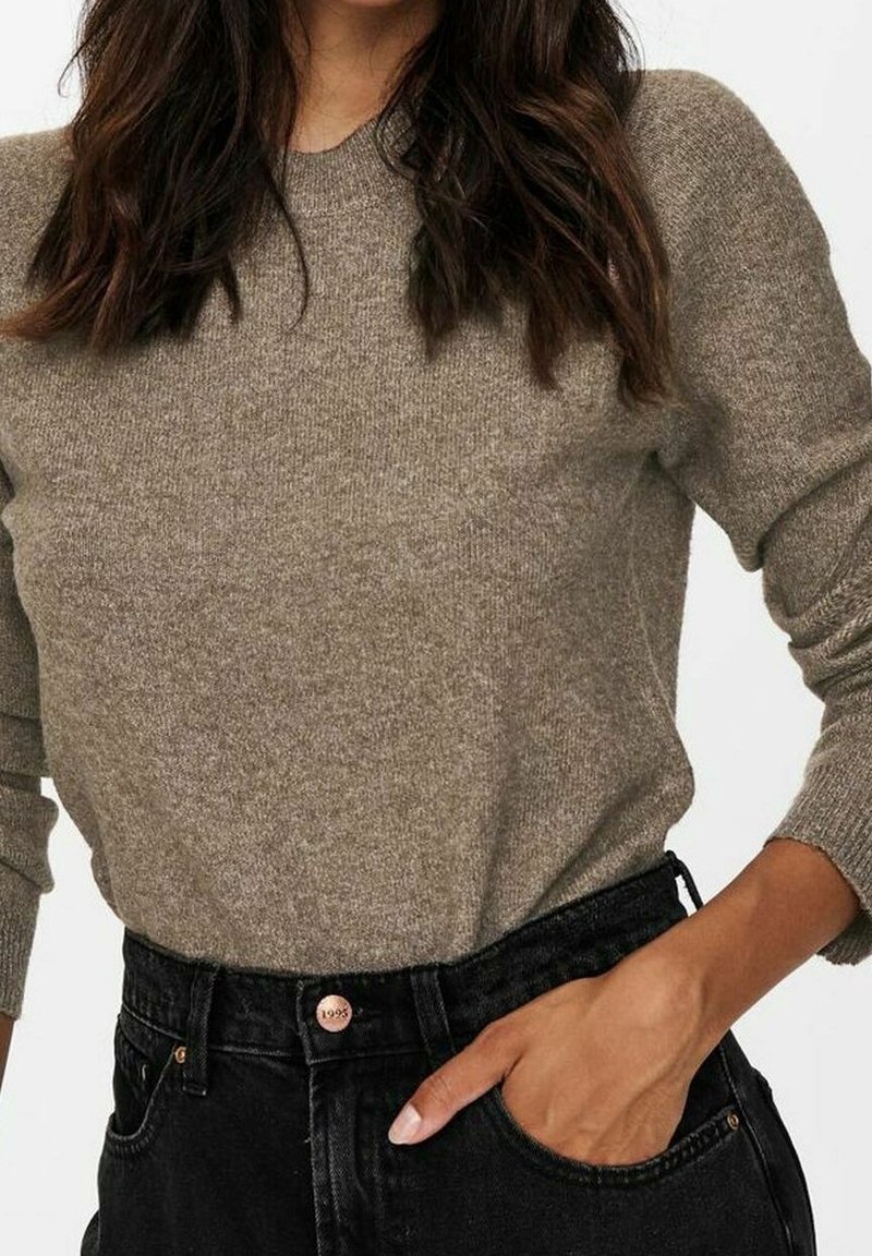 Hellbraunes Strickpullover mit gerippten Ärmeln, kombiniert mit hochgeschnittenen schwarzen Jeans, die am Bund mit einem kupferfarbenen Knopf verziert sind.
