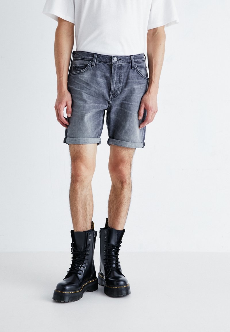Personne portant un short en denim foncé roulotté, un t-shirt blanc et des bottes militaires noires à lacets, debout sur un sol blanc uni.