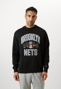 Felpa nera con la scritta "BROOKLYN NETS" e "2012" in grassetto bianco, dettagli floreali, colletto girocollo e polsini a costine, indossata con pantaloni grigi.