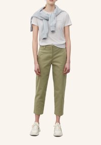 Lichtgroene, cropped broek, zachte witte t-shirt met logo en lichtblauwe trui vastgeknoopt bij de schouders. Witte sneakers maken de outfit compleet.