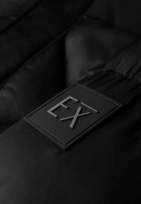 Superficie nera imbottita con un'etichetta rettangolare testurizzata in rilievo con il logo "EX". Il materiale appare lucido e liscio.