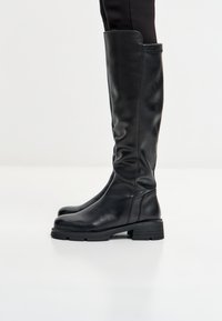 Stivali neri in pelle alti fino al ginocchio con tacco basso indossati sopra leggings neri su uno sfondo bianco semplice.