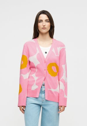 Kvinde med langt mørkt hår iført en lyserød cardigan med store gule og hvide blomsterprint over en hvid skjorte og lyseblå jeans.