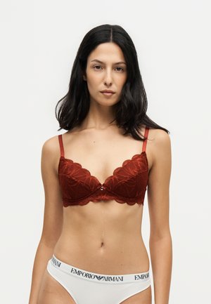 Femme aux longs cheveux foncés portant un soutien-gorge en dentelle rouge et un sous-vêtement blanc avec le logo Emporio Armani sur la ceinture, debout devant un fond uni.