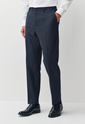 Mężczyzna ubrany w ciemnogranatowe spodnie garniturowe, czarne eleganckie buty i białą koszulę na guziki, stojący z jedną ręką w kieszeni na białym tle.