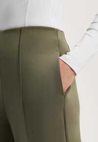 Zelene hlače iz gladkega materiala z visokim pasom, ki imajo stranske žepe in šive. Obleka je v kompletu z belo majico.