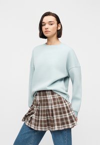 Jeune femme aux cheveux courts et foncés portant un pull oversize bleu clair, une jupe à carreaux marron superposée à un jean bleu, debout devant un fond blanc.