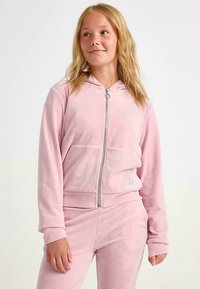 Roze hoodie met ritssluiting gemaakt van zachte stof, voorzien van een ritssluiting aan de voorkant, twee zakken en een capuchon. Soepele textuur; minimale details op de stof.