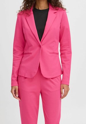 Kvinde iført en lyserød tætsiddende blazer og matchende bukser med en sort top under, stående imod en ensfarvet lys baggrund.