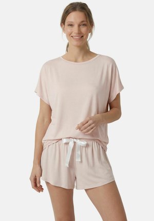 Femme souriante portant un haut rose clair à manches courtes et un short assorti avec un ruban blanc à la taille, debout devant un fond uni.