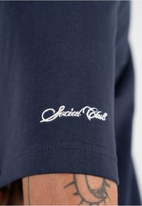 Tamsiai mėlynas medvilninis marškinėliai su trumpomis rankovėmis, su balta siuvinėta užrašu „Social Club“ ant rankovės, lygus audinys ir standartinio kirpimo.