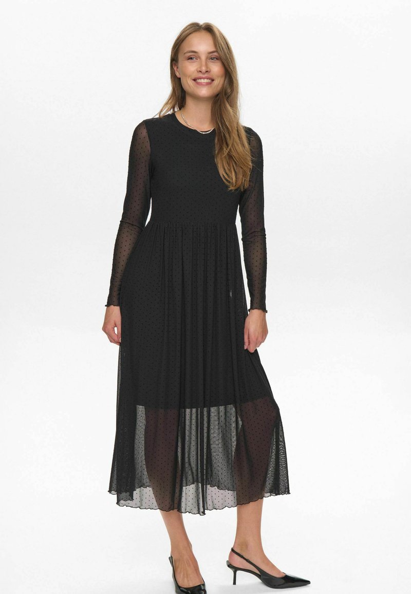 Rochie neagră cu mâneci lungi, cu un overlay transparent cu puncte, talie pliată și fustă fluidă. Asortată cu tocuri negre, având un tiv zimțat.