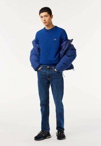 Maglione di cotone blu con scollo rotondo e dettagli verdi, abbinato a jeans blu scuro e sneakers nere, con in mano un giubbino imbottito blu navy.