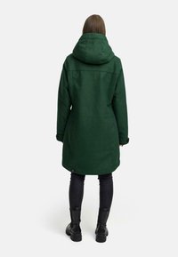 Groene lange jas met een capuchon, met een rechte snit en knoopmanchetten, gecombineerd met zwarte slim-fit broek en stevige zwarte laarzen.