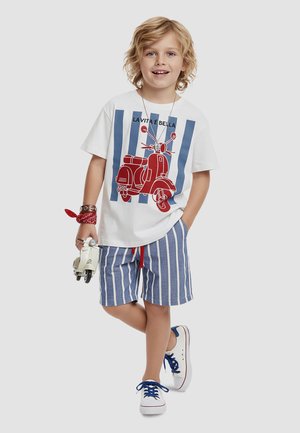 Blondes Kind trägt weißes T-Shirt mit rotem Roller-Aufdruck und blau gestreifte Shorts, hält weißen Spielzeug-Roller, lächelt, auf hellem Hintergrund.