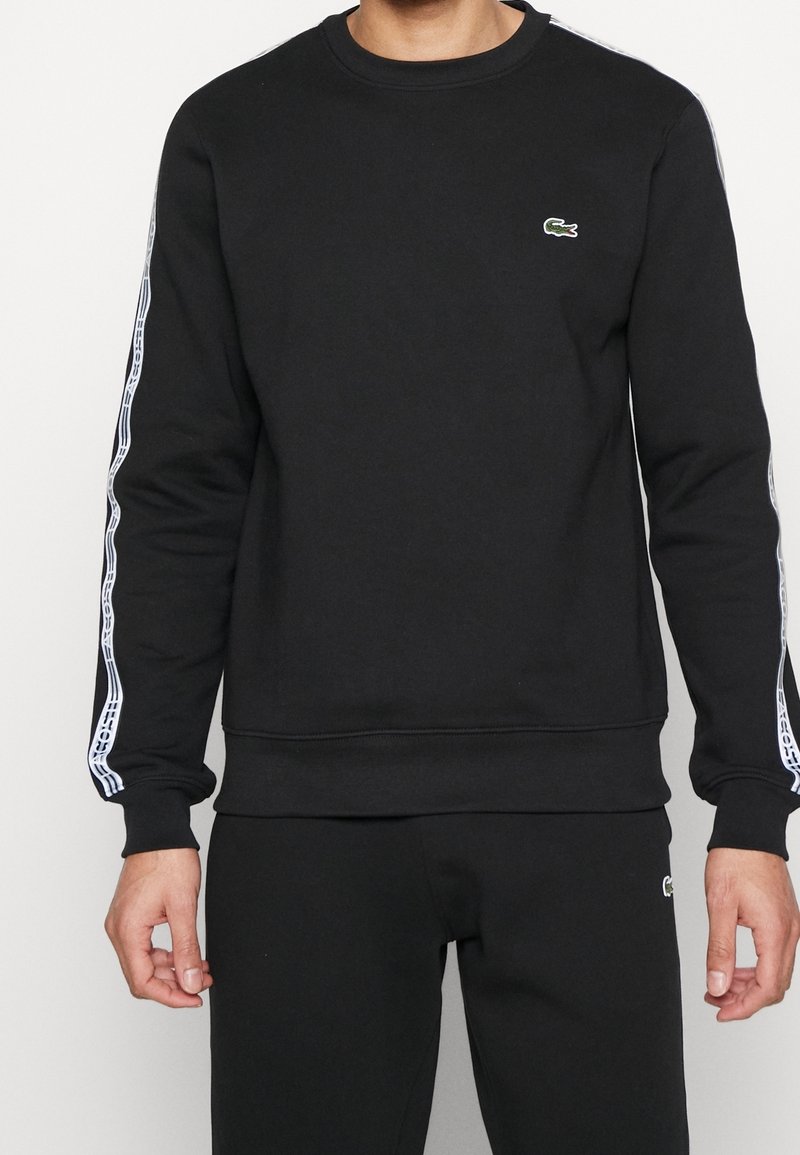 Schwarzer Sweatshirt mit Rundhalsausschnitt, gerippten Bündchen und Saum. Verfügt über weiße Seitenstreifen und ein kleines grünes Logo auf der Brust. Weicher Stoff.