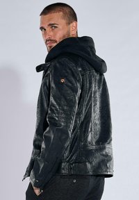 Schwarze Lederjacke mit Kapuze, mit strukturiertem Finish, Reißverschluss und seitlichen Akzenten. Besonders hervorzuheben sind die Knopf- und Logo-Details am Ärmel.