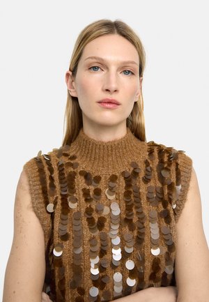 Kobieta o prostych blond włosach, ubrana w bezrękawnik z brązowego dzianinowego materiału ozdobiony dużymi błyszczącymi brązowymi cekinami, o neutralnym wyrazie twarzy.