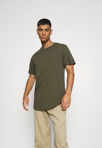Olive green short-sleeve t-shirt with a round neckline and a slightly elongated hem. Made of soft fabric, featuring minimal branding.