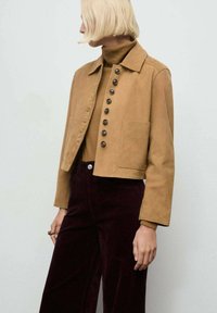 Chaqueta de ante marrón recortada con cuello y grandes botones, combinada con un suéter de cuello alto ajustado y pantalones de pana burdeos de pierna ancha.