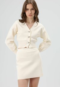 Crèmekleurige gebreide co-ord set met een cropped vest met een V-hals en drie metalen knopen, gecombineerd met een hoge taille minirok.