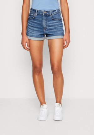 Kvinde iført mid-rise blå denimshorts med op-rullede kanter, lyseblå top og hvide sneakers, der står mod en ensfarvet hvid baggrund.