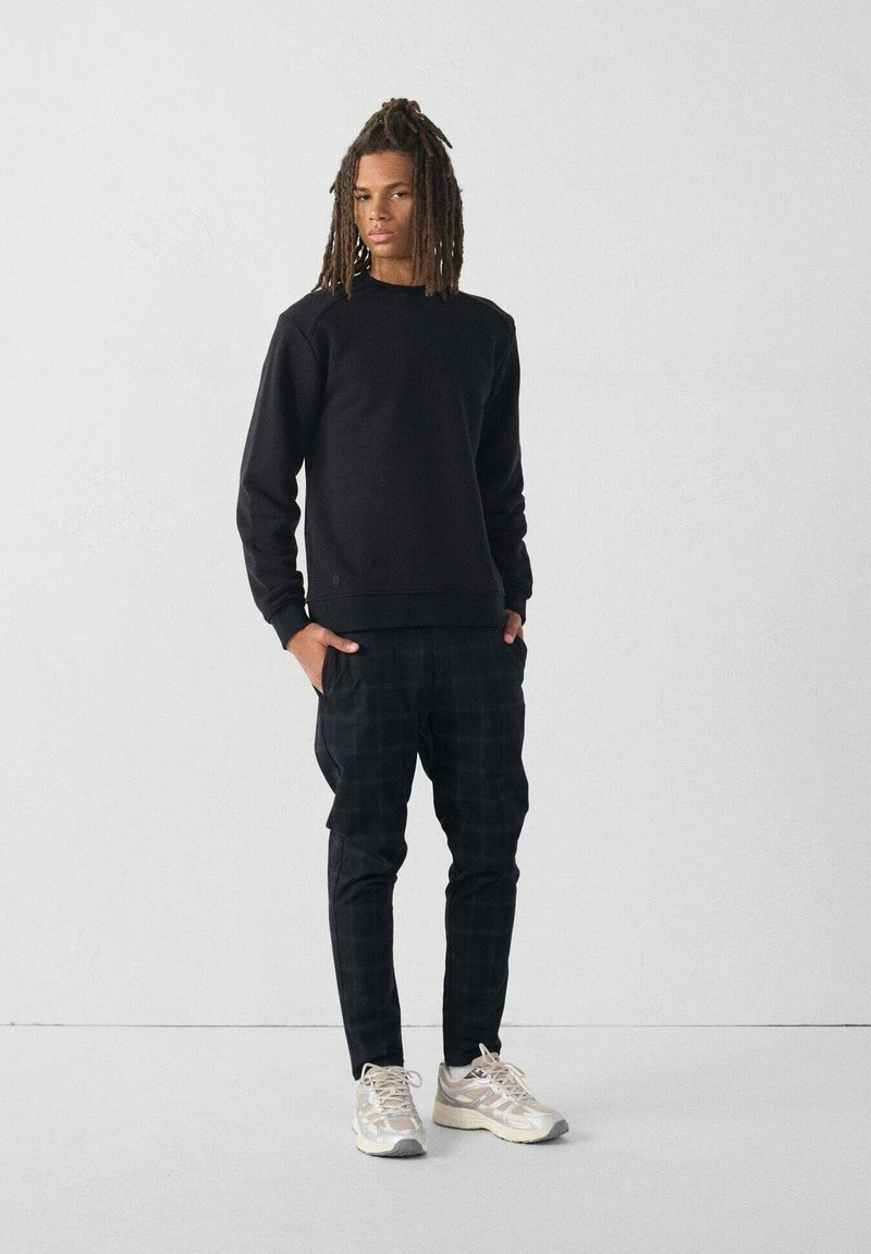 Sweatshirt noir à col ras du cou associé à un pantalon à carreaux noir. Le mannequin porte des baskets claires avec des accents réfléchissants. Design simple et décontracté.