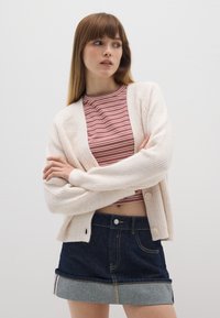 Hellbeige gestrickte Strickjacke mit gerippter Textur und vorderen Knöpfen, getragen über einem rot-weiß gestreiften Crop-Top, kombiniert mit einem dunklen Jeansrock.