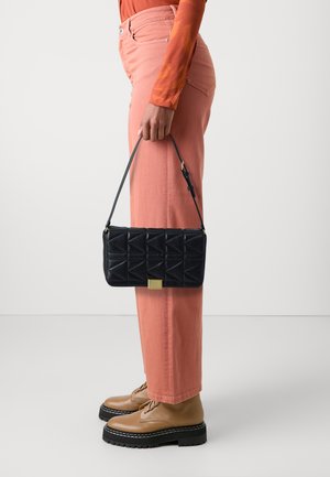 Black quilted handbag with geometric design, gold hardware, and a short strap, held alongside pink wide-leg pants and brown lace-up boots.