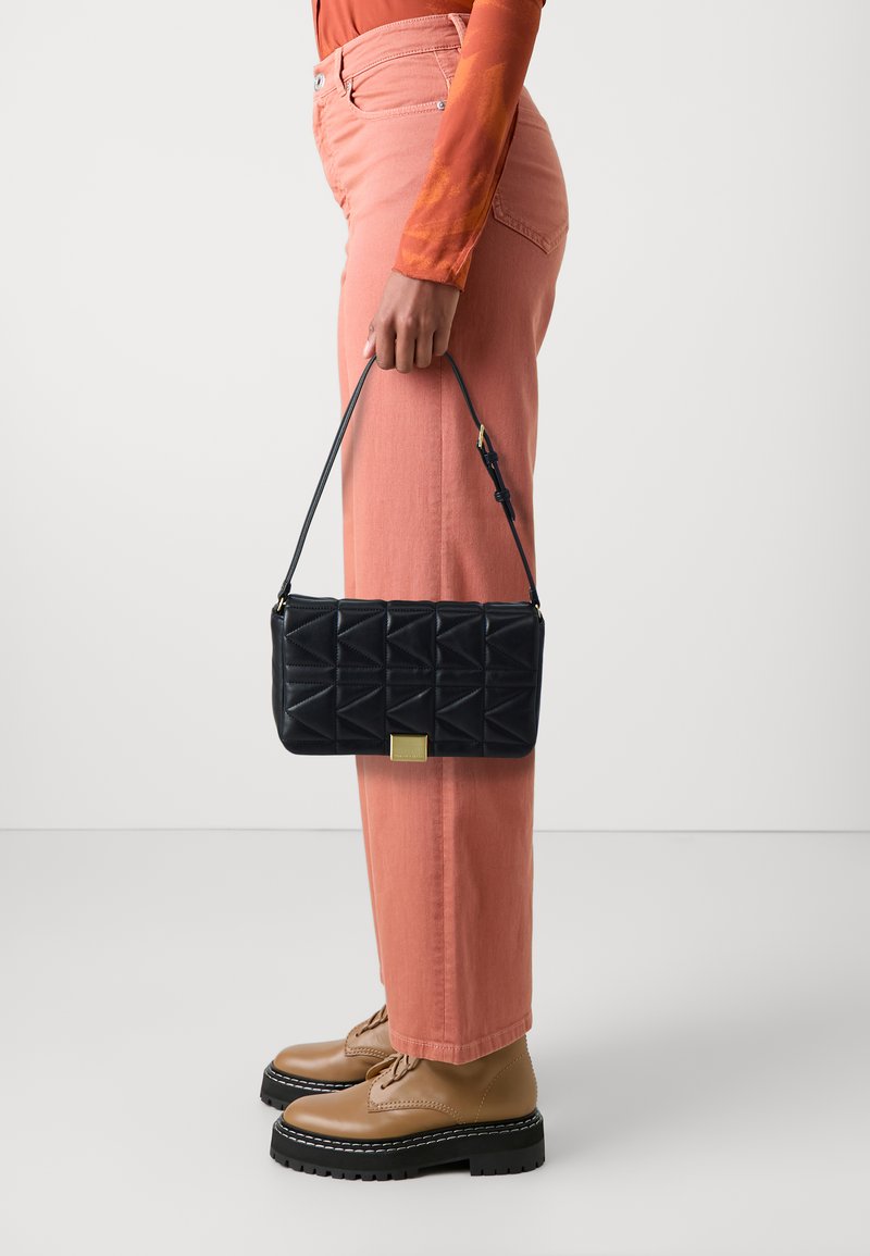 Sac à main noir matelassé avec un design géométrique, quincaillerie dorée et une courte bandoulière, tenu à côté d'un pantalon large rose et de bottes brunes à lacets.