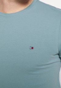 Světle modré bavlněné tričko s kulatým výstřihem a malým vyšitým logem v červené, bílé a modré barvě na levé hrudi.