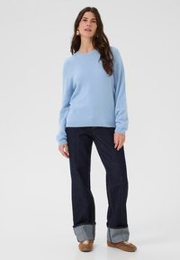 Egy nő áll, világoskék pulóverben, sötét, felhajtott szárú széles szárú farmert visel, barna tornacipőt és arany karikafülbevalót hord, síma háttér előtt.