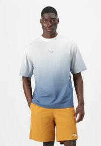 Ombre t-shirt in white to blue gradient, featuring a logo near the collar. Paired with mustard yellow shorts made from lightweight fabric.