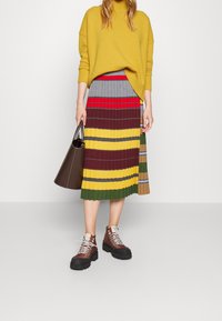 Kobieta w żółtym swetrze i kolorowej plisowanej spódnicy, trzymająca brązową torebkę i nosząca brązowe buty na sznurówki.