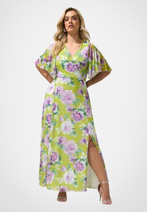 Femme portant une longue robe fleurie vert citron avec des fleurs violettes, des manches volantées, un col en V et une fente haute à la cuisse, associée à des talons argentés.