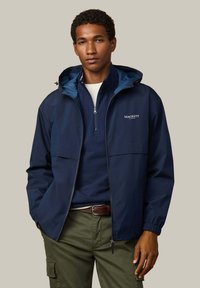 Námornícka bunda s kapucňou, so zapínaním na zips, dvoma prednými vreckami a elastickými manžetami. Nosená cez námornícky top a olivovo zelené nohavice.