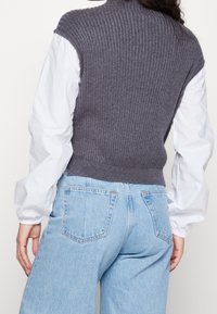 Grauer gestrickter Pullover mit gerippter Textur, hohem Kragen und kurzen Ärmeln, getragen über einem weißen Langarmshirt und kombiniert mit hellblauer Denim.