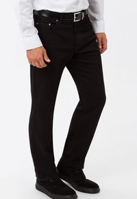 Hombre vestido con jeans ajustados negros, cinturón negro, camisa blanca y zapatos de ante negros, de pie frente a un fondo blanco.