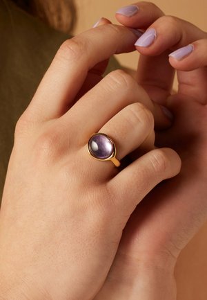 Bague en or avec une pierre ovale de couleur violet, affichant une texture lisse et une surface brillante, portée sur une main à la peau claire et aux ongles lavande.