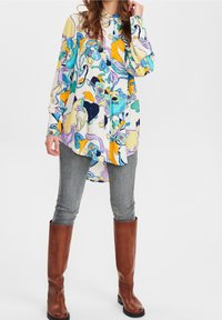 Camisa larga de mangas largas y colores vivos con un estampado floral en azules, amarillos y verdes, combinada con jeans ajustados grises y botas marrones hasta la rodilla.