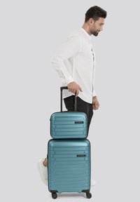 Set di valigie rigide turchesi impilate; valigia grande con righe orizzontali e quattro ruote, borsa di dimensioni cabine più piccola con design abbinato.