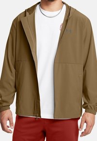 Veste légère marron avec fermeture éclair sur le devant, poches latérales et poignets élastiques, portée sur un T-shirt blanc avec inscription rouge.