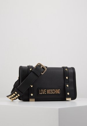 Czarna torebka na ramię Moschino z złotymi ćwiekami, regulowanym paskiem i złotą tabliczką z logo na przodzie, przedstawiona na białym tle.