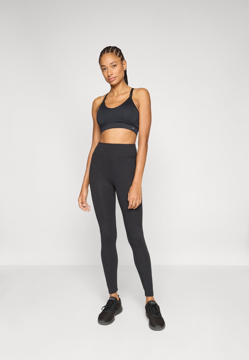 Sort sports-bh og højtaljede leggings lavet af strækbart, glat stof. Model iført sorte sports-sko, stående mod en neutral baggrund.