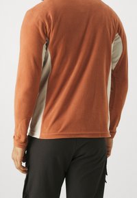 Polarowy pulower z długimi rękawami w kolorze przypalanej pomarańczy i beżu. Charakteryzuje się miękką teksturą, kontrastującymi panelami bocznymi oraz zaokrąglonym dołem.