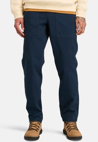 Navy blue, relaxed-fit cargo pants made of durable cotton with two front pockets; paired with tan hiking boots, featuring lace-up design.