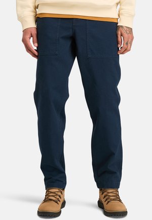 Pantaloni cargo blu navy, dal taglio rilassato, realizzati in cotone resistente con due tasche frontali; abbinati a scarponi da trekking color sabbia, con design stringato.